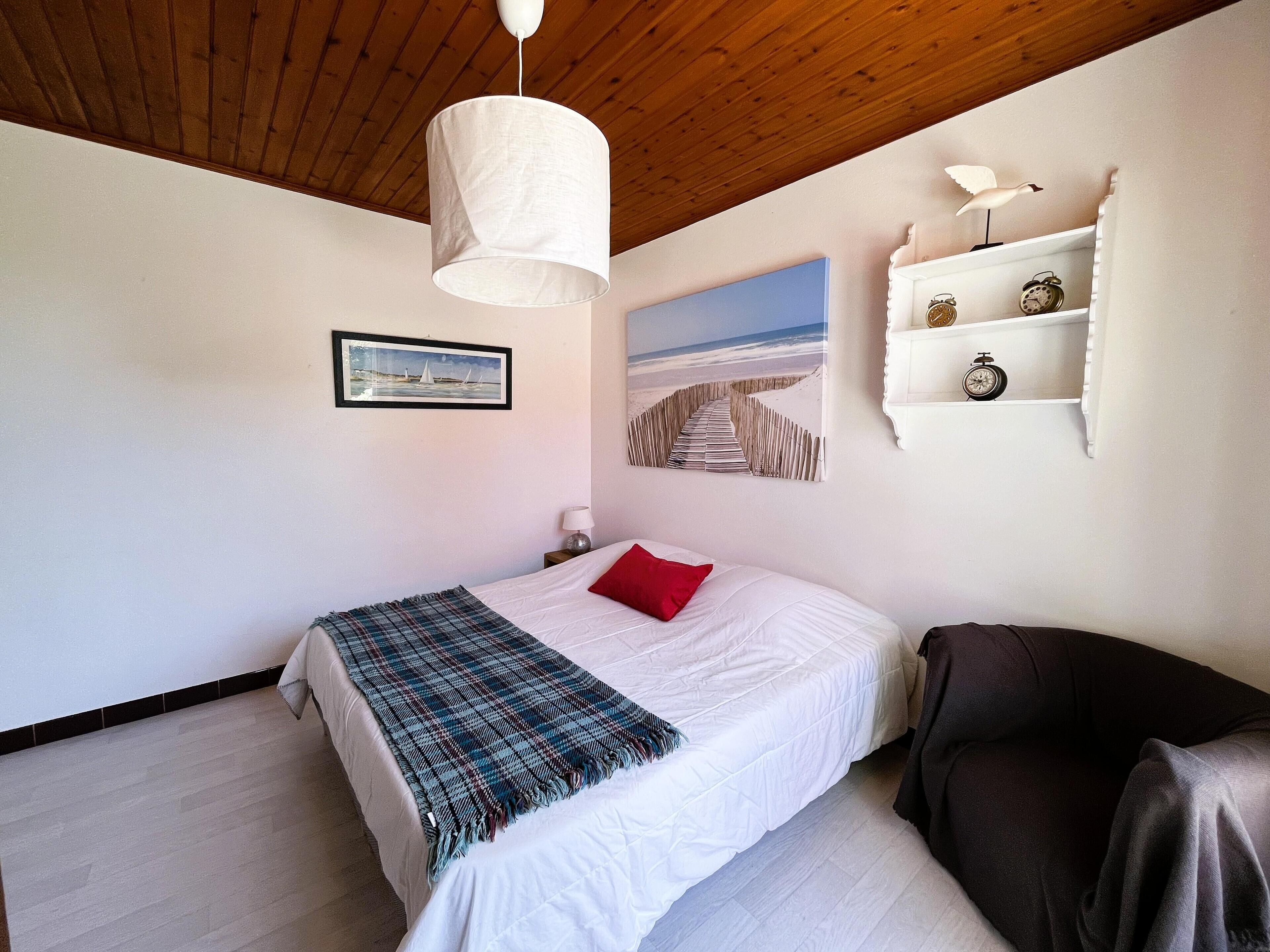 A bedroom with a bed, a wooden ceiling, a white wall, a shelf with decorative items, and a painting of a beach scene.