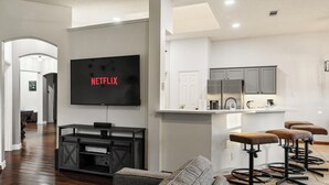 A modern living room with a flat-screen TV displaying "NETFLIX," a kitchen with grey cabinets, and a bar area with stools. - 2 minutes from lake, Pickleball, Hot Tub, Pool table! (Rowlett)