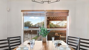 A dining area with a wooden table set for four, a potted plant, and a view of the outside through large windows. - 2 minutes from lake, Pickleball, Hot Tub, Pool table! (Rowlett)