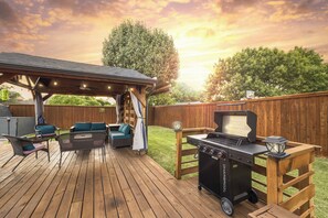 Una terraza de madera con mobiliario exterior, una parrilla y un área de estar cubierta.