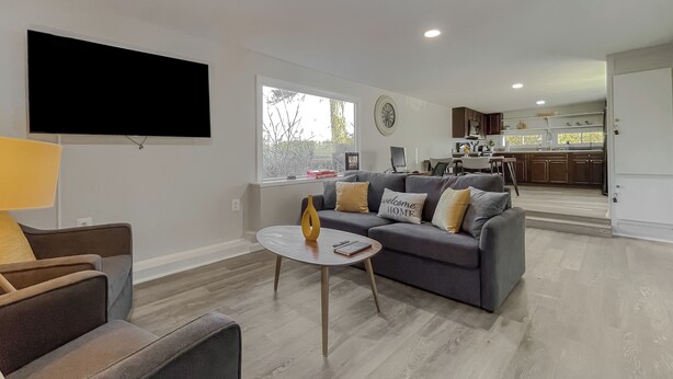 A modern living room with a grey sofa, a coffee table, and a TV mounted on the wall. A kitchen is visible in the background with wooden cabinets and a window.