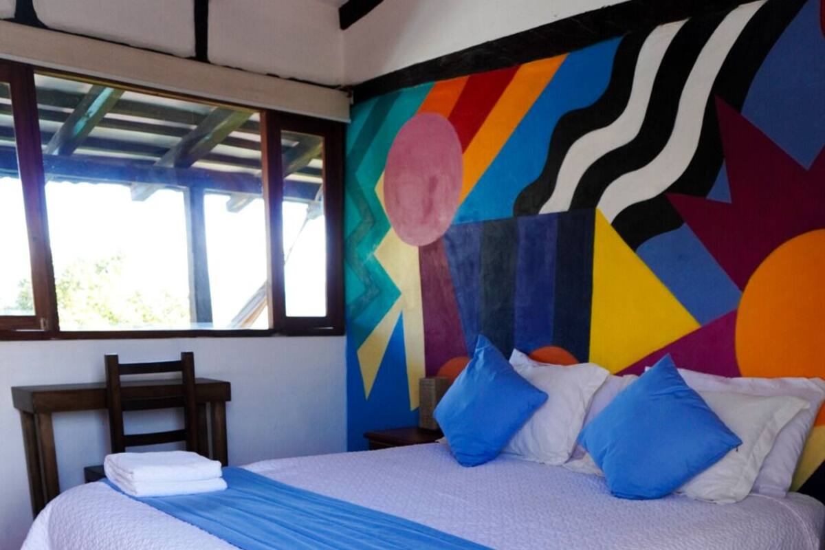 Un lit avec une couvre-lit blanche et bleue, une tête de lit en bois et une fresque murale abstraite et colorée.