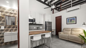 Une cuisine moderne avec des armoires blanches, un comptoir en bois et un réfrigérateur en acier inoxydable. Le salon est doté d’un canapé beige et d’un ventilateur de plafond.