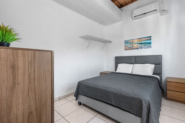 A modern bedroom with a grey bed, a wooden wardrobe, a wall-mounted air conditioner, a small bedside table, and a painting of a sunset. - Beach Access Escape With Pool and Tanning Lounges (San Juan)