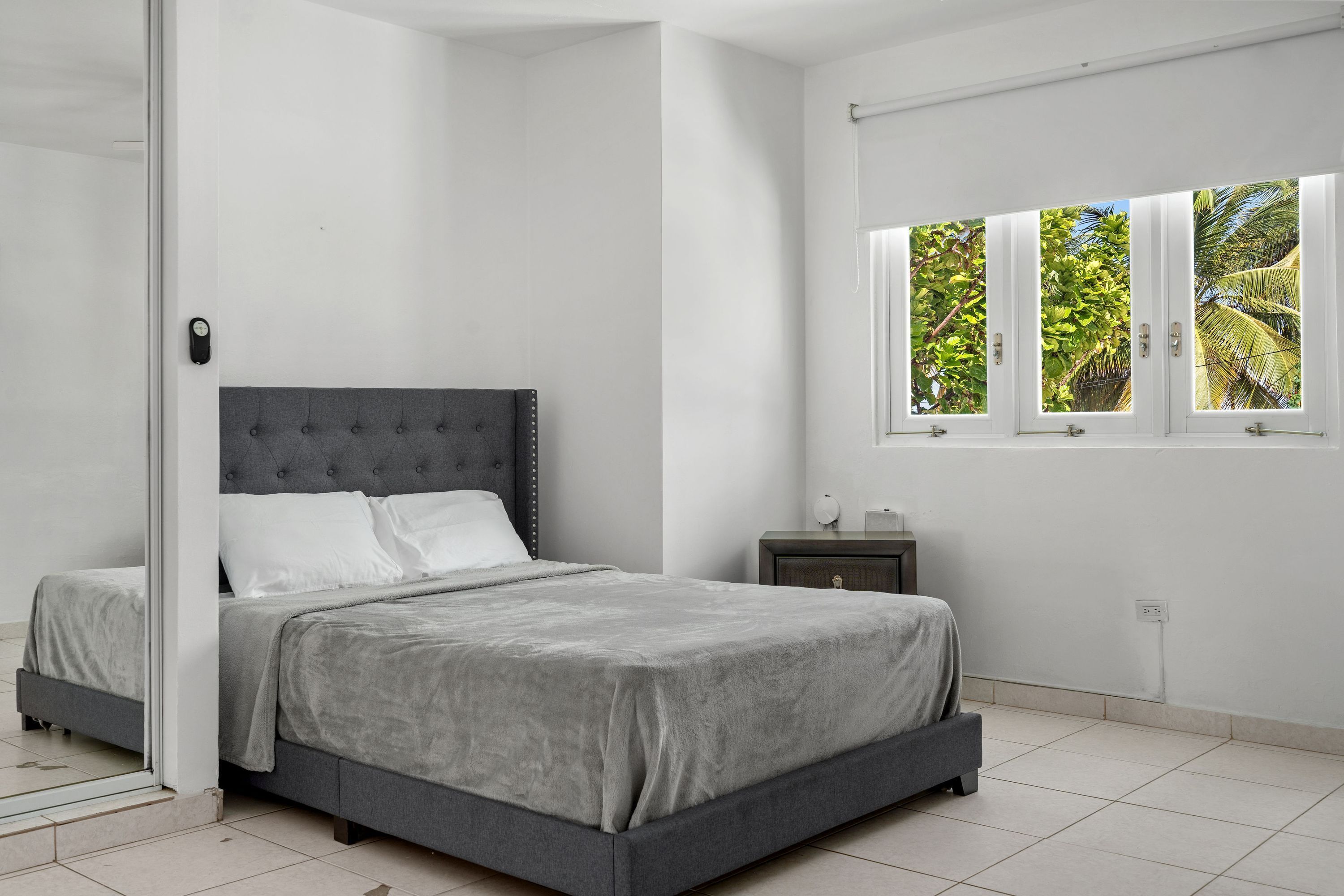 Une chambre à coucher comprenant un lit, une table de chevet, une fenêtre donnant sur un espace verdoyant et une porte.