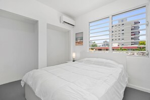 A minimalist bedroom with a large bed, an air conditioning unit, a bedside lamp, and a window with a view of buildings and greenery. - Otium Walk to the Beach Free Local Coffee (San Juan)