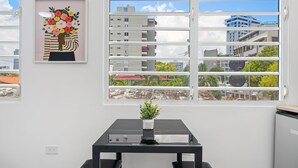 Une pièce avec une grande fenêtre, une œuvre d’art encadrée, une petite table sur laquelle est posée une plante en pot et une chaise noire.