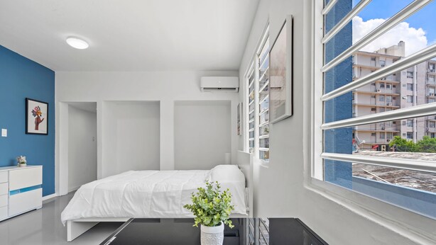 Une chambre à coucher moderne, avec un lit, une table de chevet, une plante en pot et une fenêtre donnant sur des immeubles.
