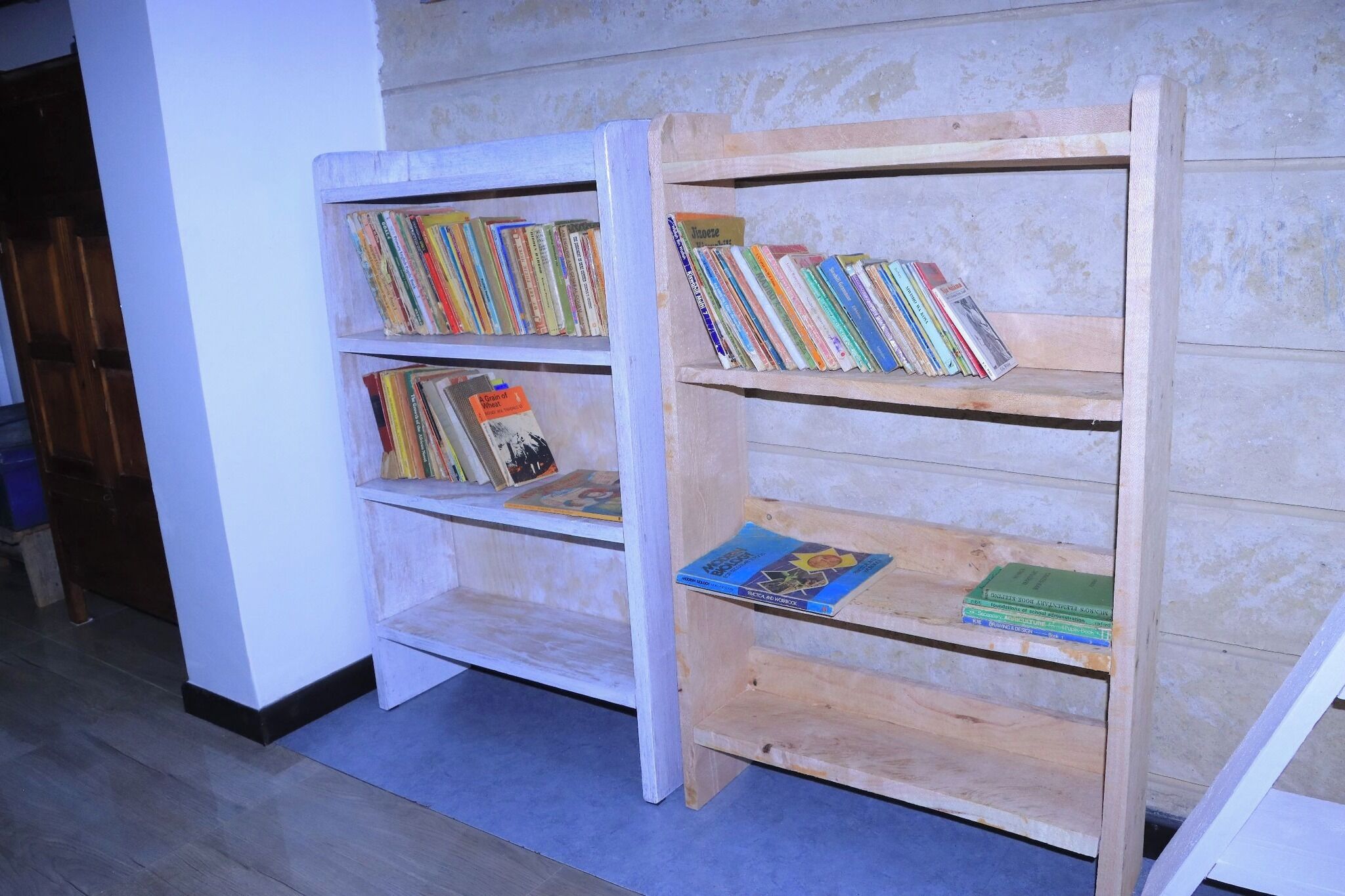 Deux étagères en bois remplies de livres, l’une adossée à un mur et l’autre à un tableau blanc.