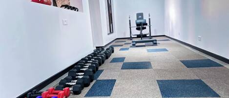 A hallway with a row of dumbbells on the left and a weight bench in the distance.