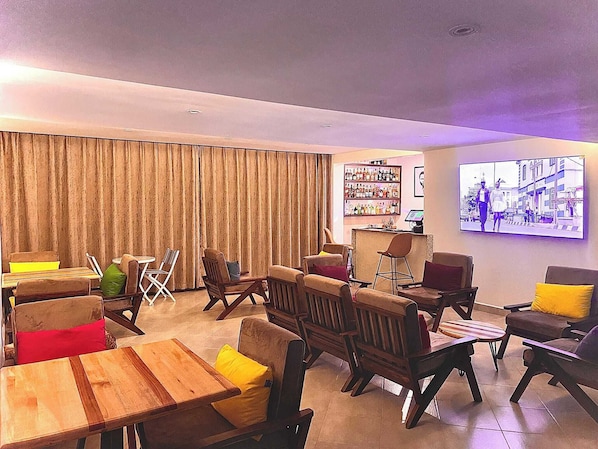 A modern lounge area with wooden furniture, a bar, and a large TV screen displaying a cityscape. - Humphreys Hill House (Kisii)