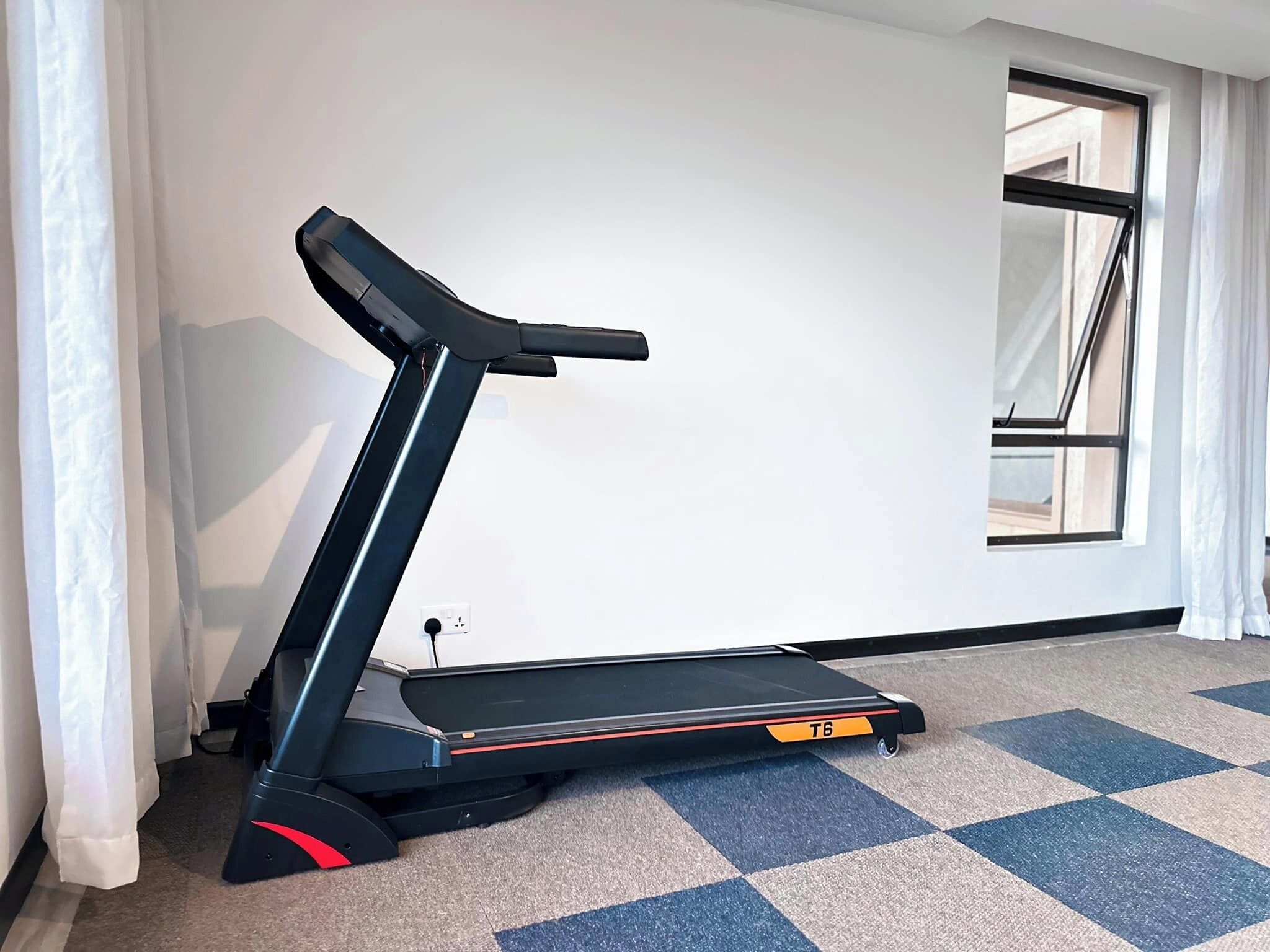A treadmill in a room with a checkered carpet, a window with curtains, and a white wall.