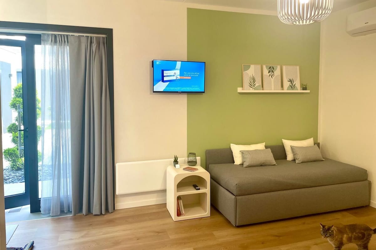 A modern living room with a grey sofa, a small white side table, a TV mounted on the wall, and a green accent wall.