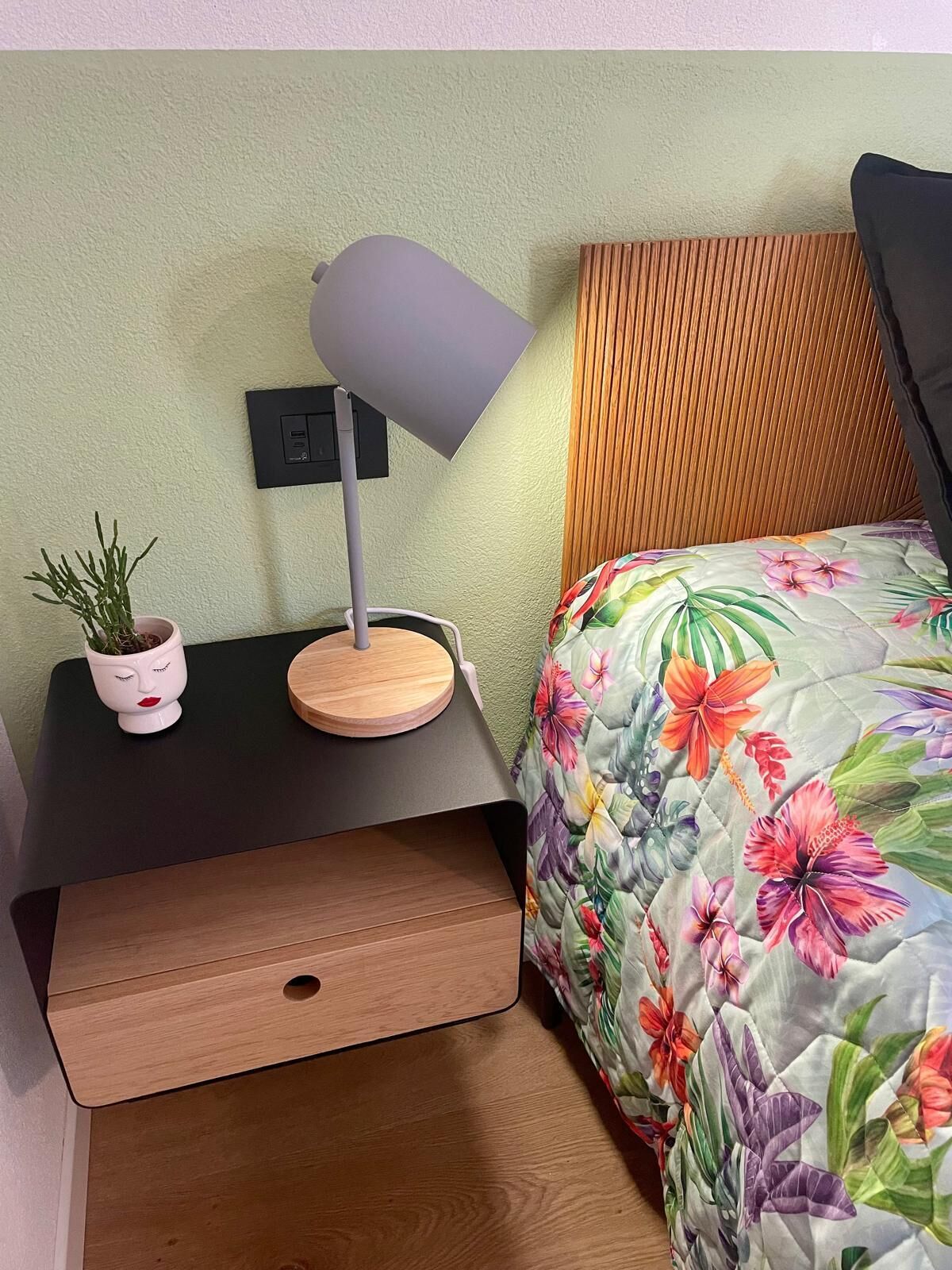 A neatly arranged bedroom with a floral bedspread, a bedside table with a lamp, and a small potted plant.