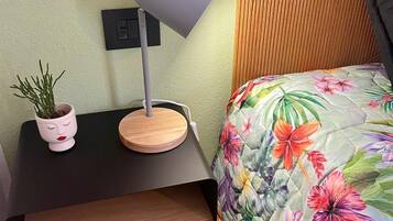 A neatly arranged bedroom with a floral bedspread, a bedside table with a lamp, and a small potted plant.