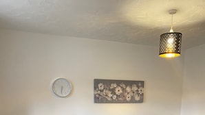 A living room with a black sectional sofa, a wooden dining table with chairs, a patterned rug, a wall clock, and a floral painting.