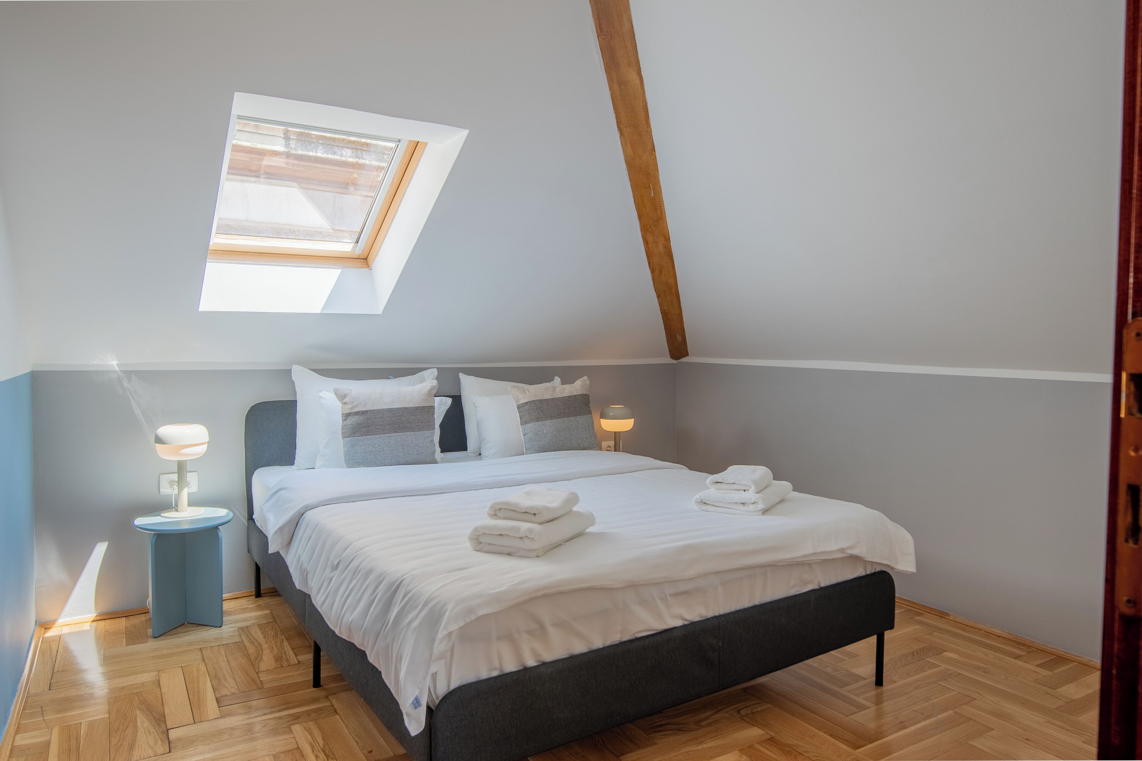 A bedroom with a skylight, a bed with white linens, two bedside tables with lamps, and a herringbone-patterned floor.
