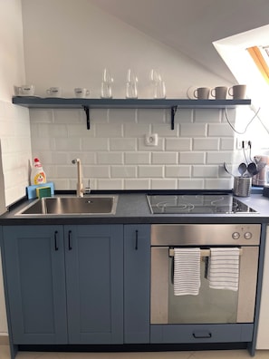 Ett modernt kök med en diskho i rostfritt stål, en inbyggd spis och en mikrovågsugn. Köket har skåp med handtag, en hylla med koppar och glas, och ett fönster ovanför diskhon.