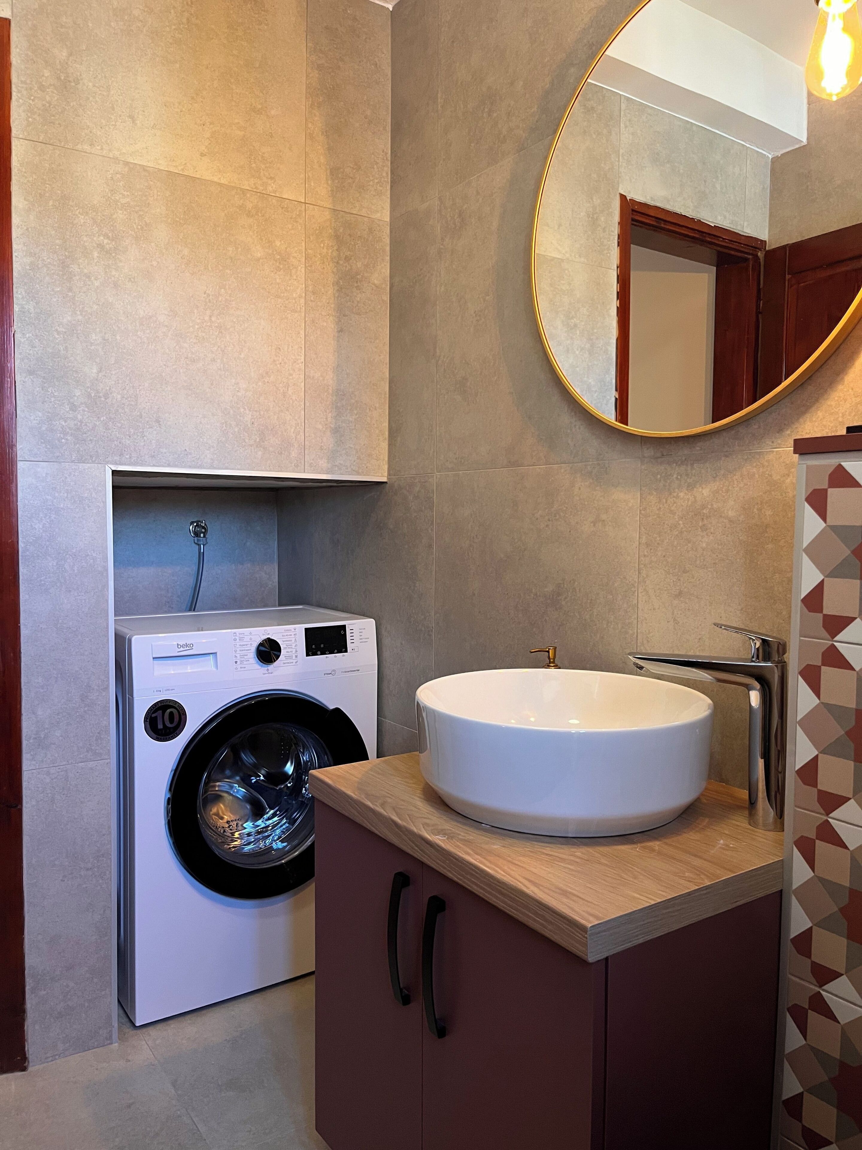 A modern bathroom with a built-in washing machine, a round mirror, and a sink with a chrome faucet.