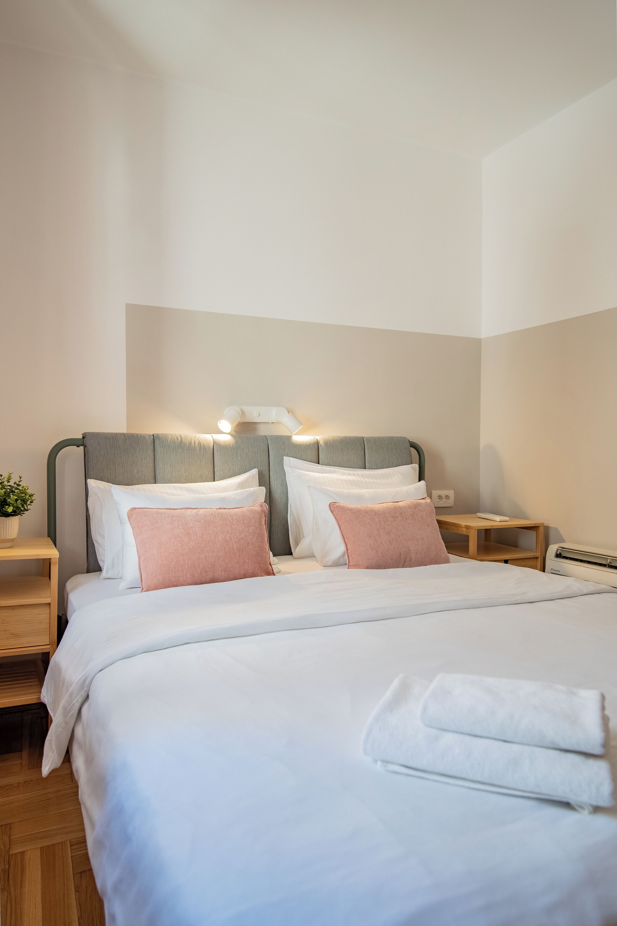 Ein ordentlich bezogenes Bett mit weißen und rosafarbenen Kissen, einer Nachttischlampe und einem kleinen Tisch mit einer Pflanze.