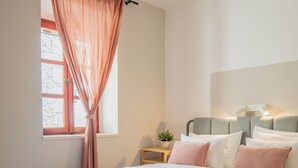 A bedroom with a bed, a window with curtains, a bedside table with a lamp, and a potted plant. - Old Town Church Bells (Kotor)