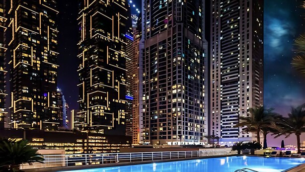 Ein Nachtbild eines modernen Hochhauskomplexes mit beleuchteten Fenstern und einem Schwimmbecken im Vordergrund.