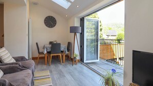 Ein modernes Wohnzimmer mit einer Couch, einem Esstisch mit Stühlen, einer Stehlampe, einem Fernseher und einem Balkon mit Blick auf Grünflächen.