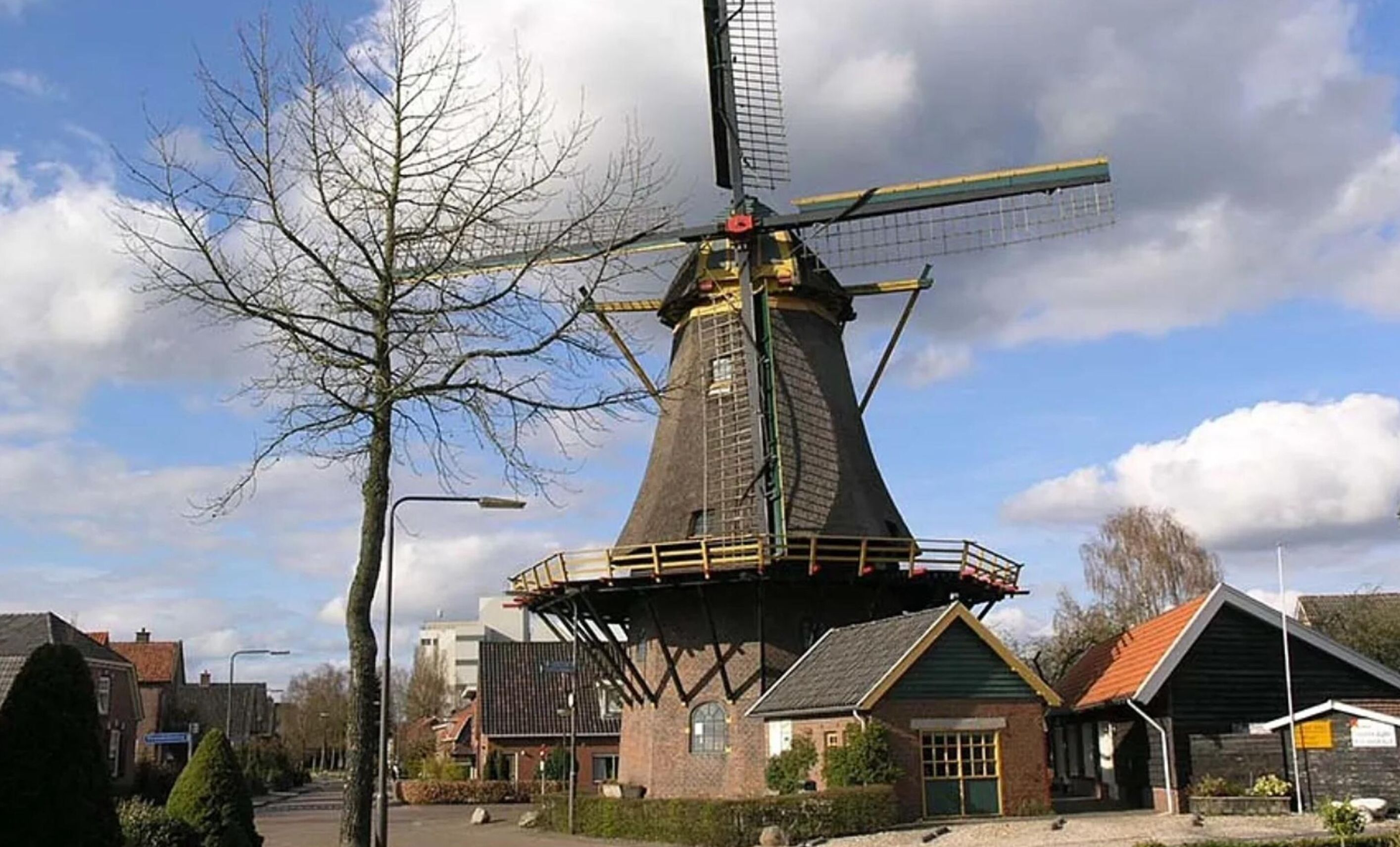 A traditional windmill with a conical roof and a large sail, surrounded by residential buildings and a leafless tree.