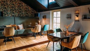 Un espace repas doté d’un plancher en bois, d’un plafond haut et d’une grande fenêtre. On y trouve des chaises et une table sur laquelle est posé un vase rempli de fleurs.