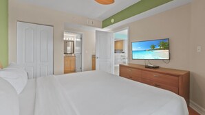 Une chambre d’hôtel avec un lit, une commode, un téléviseur diffusant une scène de plage et, au fond, une petite cuisine.