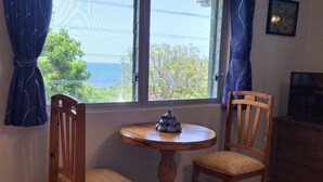A room with a round wooden table and two wooden chairs with cushioned seats. A window with curtains is visible, and a painting hangs on the wall.
