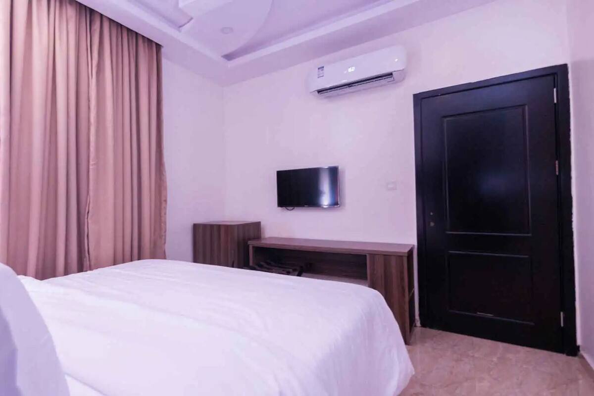 Une chambre d’hôtel avec un lit, un téléviseur à écran plat fixé au mur, un bureau en bois et une porte noire.