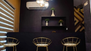 A modern interior with a black wall, a white air conditioner, a decorative light fixture, a small shelf with plants, and three gold wire chairs. - Ibizza Pit Hotel and Suites (Lagos)