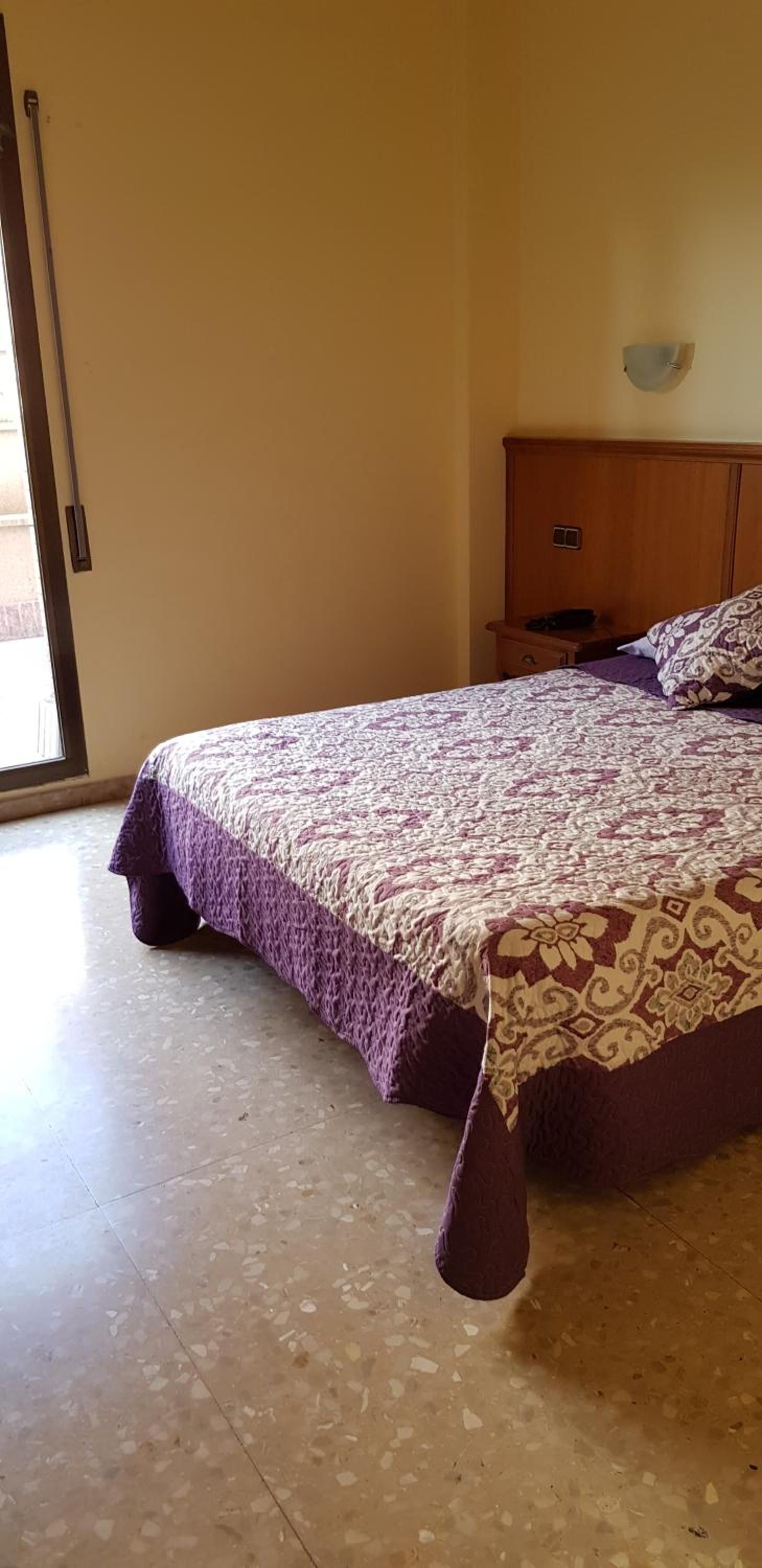 A bedroom with a bed, a wooden nightstand, a wall-mounted light, and a sliding glass door.