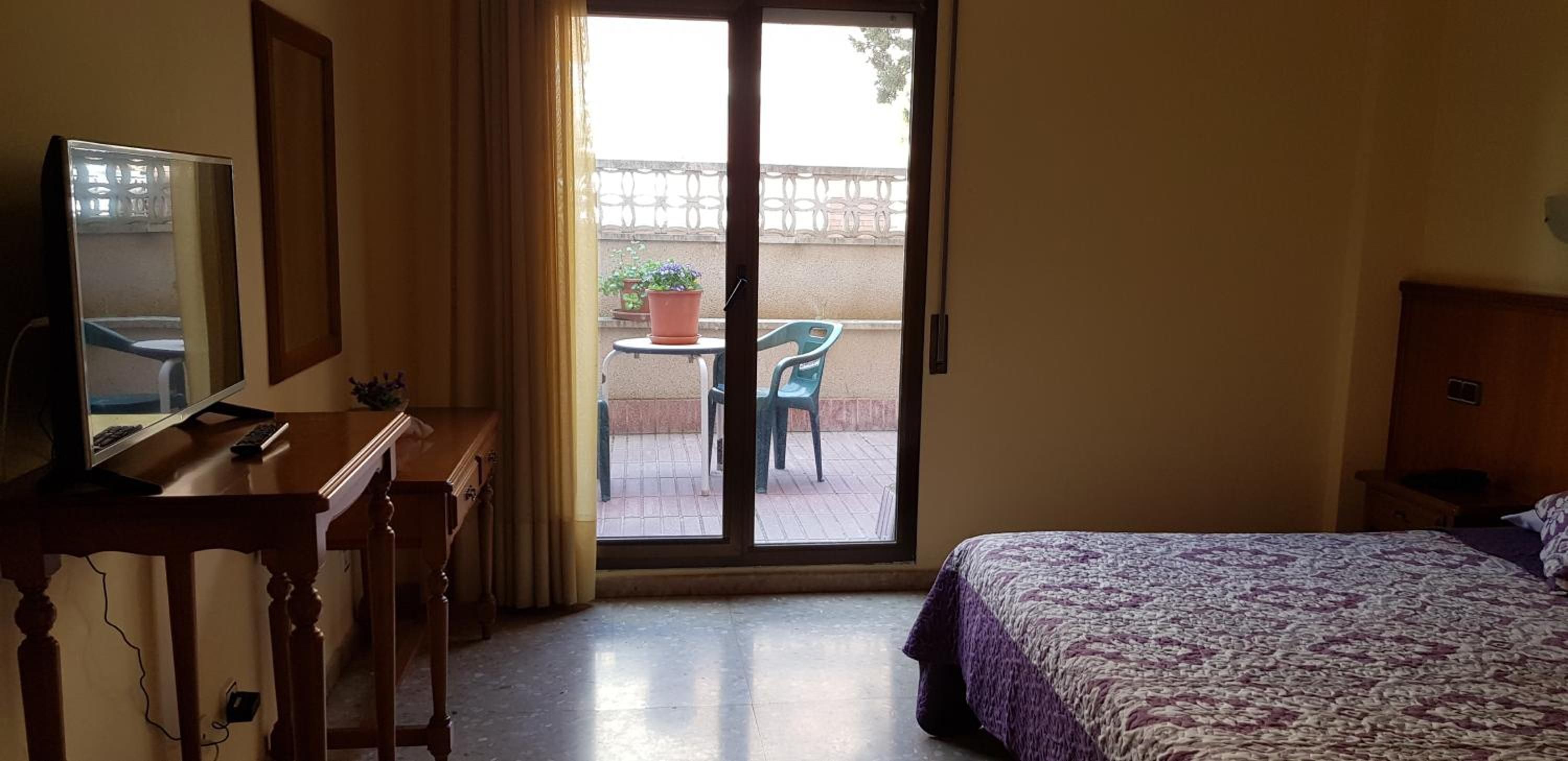 A hotel room with a bed, a desk with a computer, a chair, and a balcony with a table and chairs.