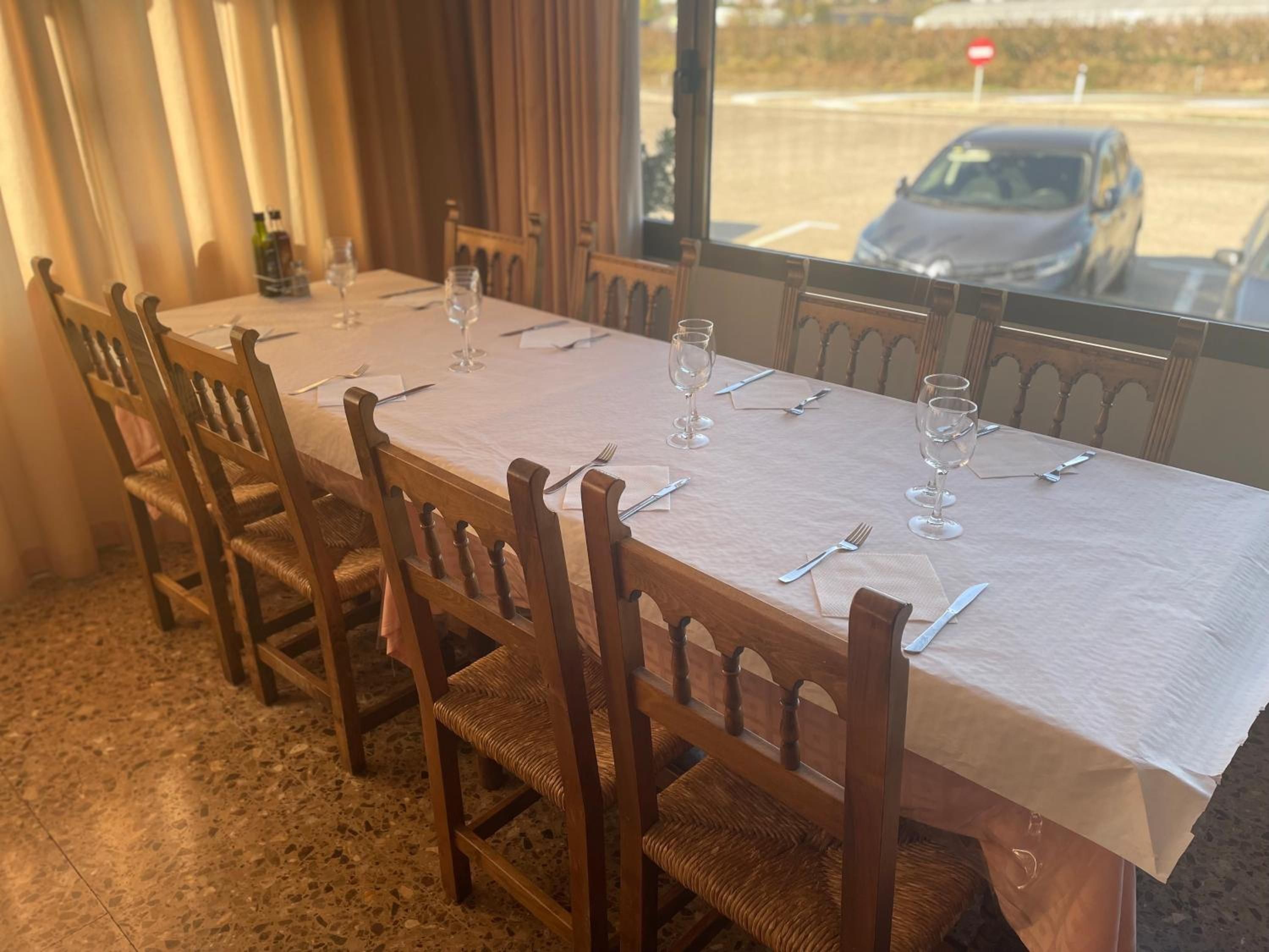 Una mesa de comedor con sillas, vista por la ventana a un estacionamiento y una puerta con cortinas.