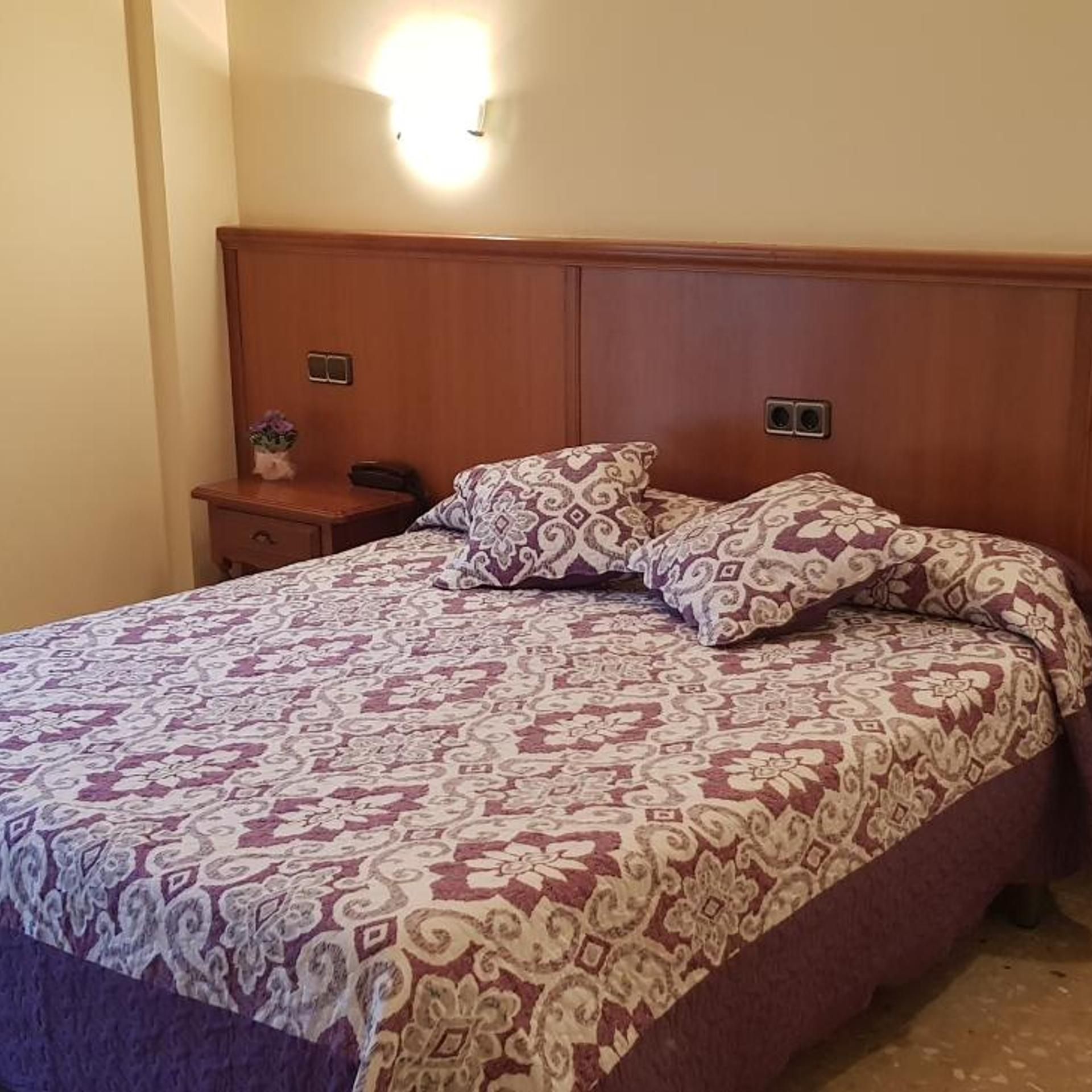 A bed with a patterned bedspread and two pillows, a bedside table with a lamp, and a wall-mounted light fixture.