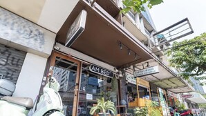 Une devanture de magasin avec une enseigne indiquant « AM BED » et une autre enseigne dans une langue différente. Un scooter est garé devant, et divers pots de plantes et objets sont exposés à l’extérieur.