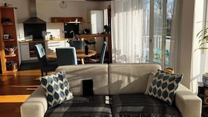 Una sala moderna con un sofá, dos cojines estampados, un control remoto y un televisor. Se ve un área de comedor con una mesa y sillas al fondo.