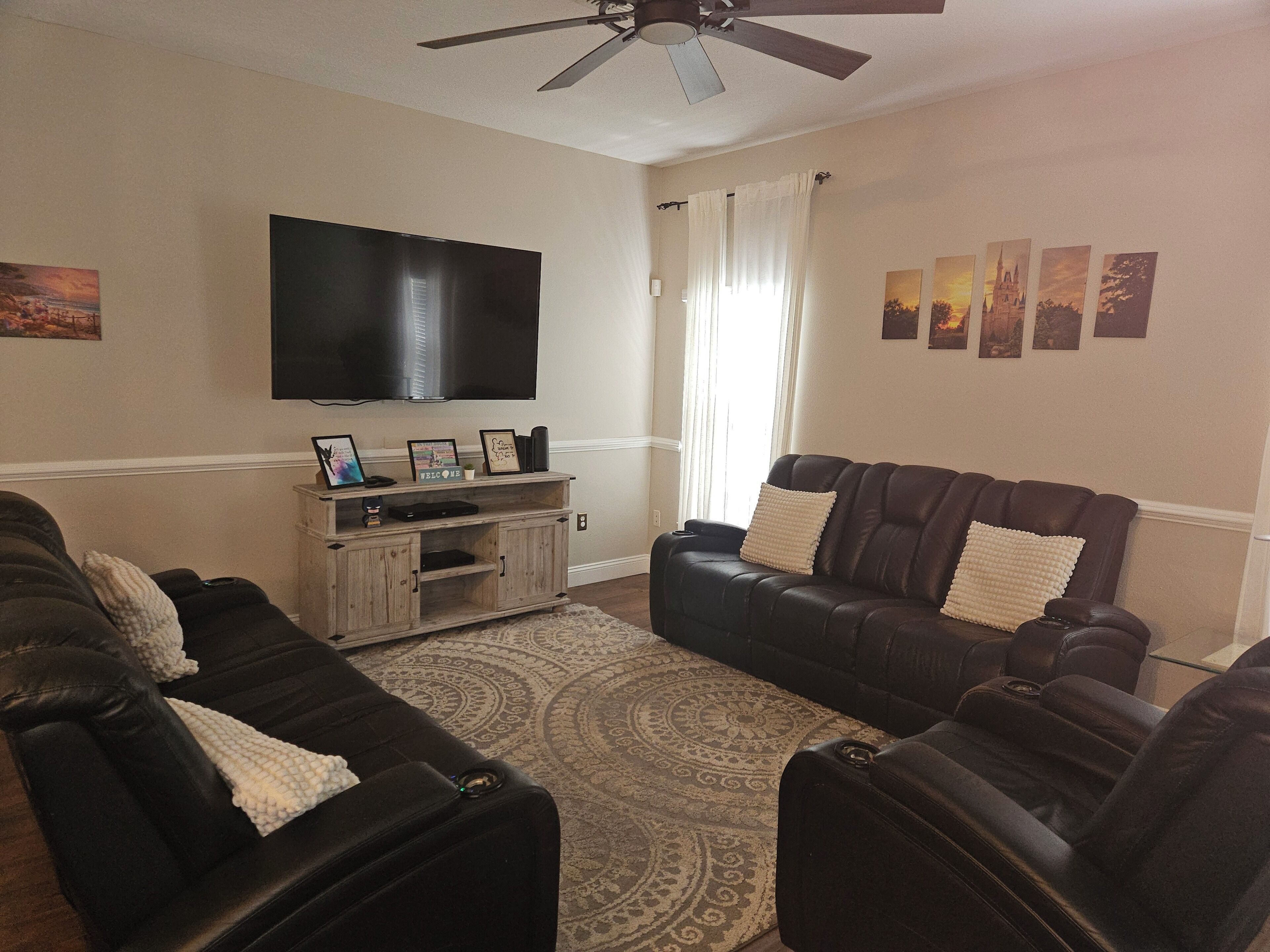 Una sala de estar con televisor de pantalla plana, un mueble bajo para equipo audiovisual de madera, dos sofás de cuero, un tapete estampado y un ventilador de techo.
