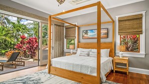 A four-poster bed with a wooden frame, a large window with a view of tropical plants, and a small bedside table with a lamp.