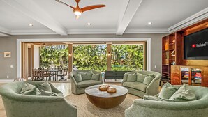 A spacious living room with a large TV, green sofas, a wooden coffee table, and a ceiling fan.