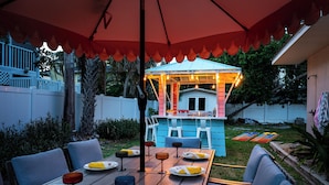 An outdoor dining area with a table set for four, surrounded by chairs, under a large umbrella. - 3 Blocks to Beach ☀️ Game room, cabana bar + family fun (Holmes Beach)