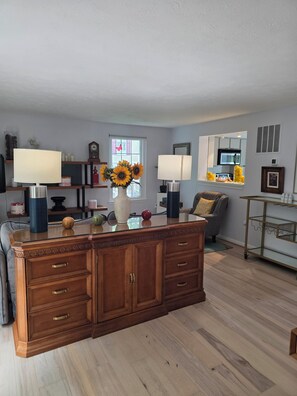 A living room with a wooden cabinet, two lamps, a vase with sunflowers, a sofa, and a kitchen area in the background.