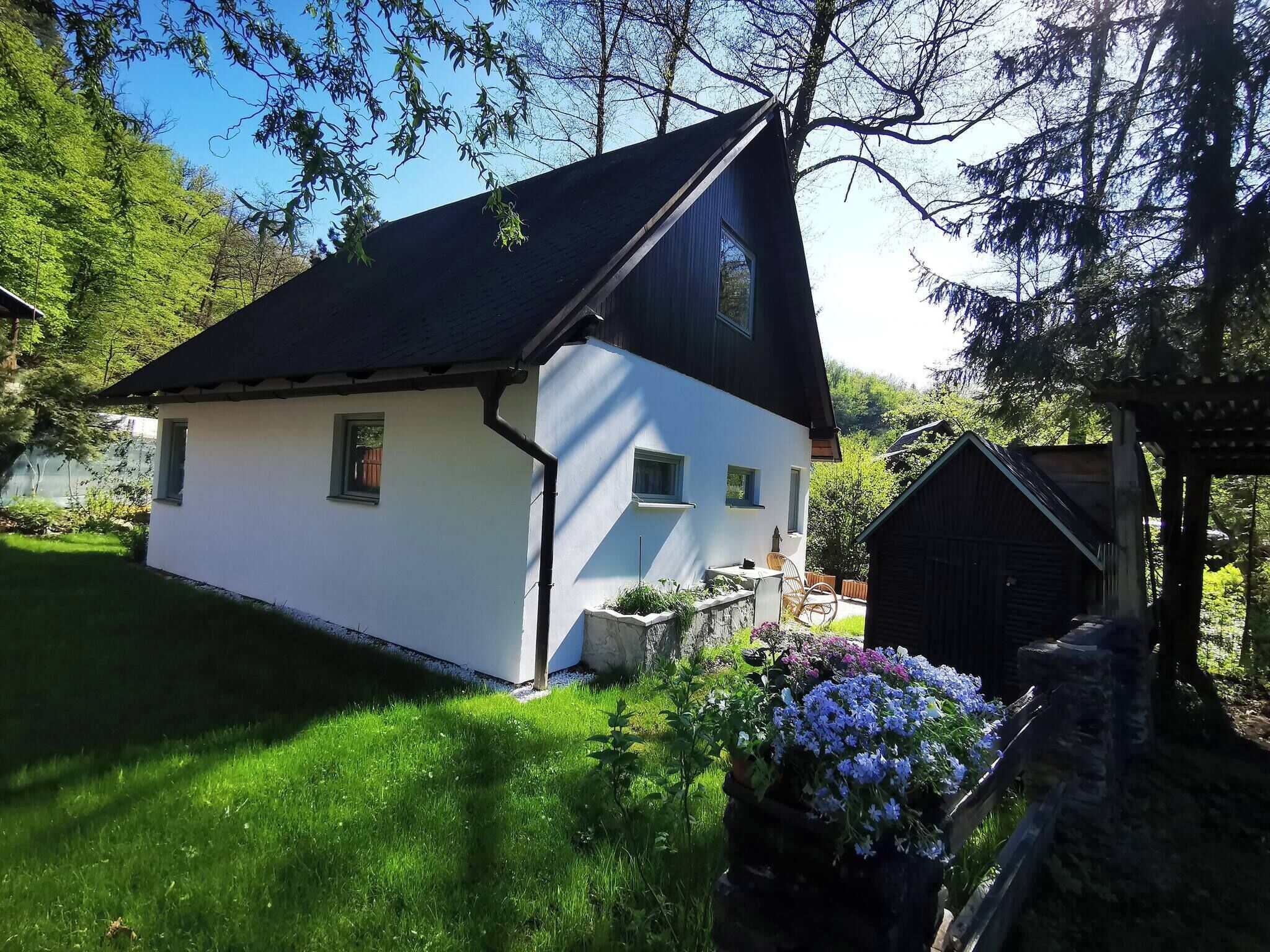 A white house with a black roof, surrounded by a garden with flowers and a small wooden structure.