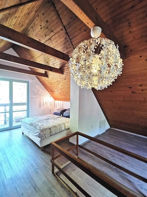 Une chambre avec un plafond en pente, des poutres en bois, un lit recouvert d’une couvre-lit à motifs floraux, un luminaire suspendu et une grande fenêtre.