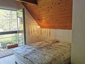 Un dormitorio con techo inclinado de madera, una cama con ropa de cama floral y vistas a la vegetación a través de un amplio ventanal.