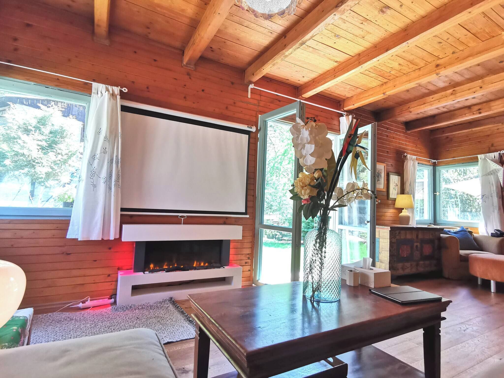 A cozy cabin interior with a fireplace, wooden walls, a large window with curtains, a table with a vase of flowers, and a couch.