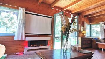 A cozy cabin interior with a fireplace, wooden walls, a large window with curtains, a table with a vase of flowers, and a couch.