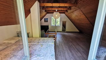 Amplia habitación en ático con vigas de madera, cama con ropa de cama floral, un banco de madera y ventana con vistas a zonas verdes.